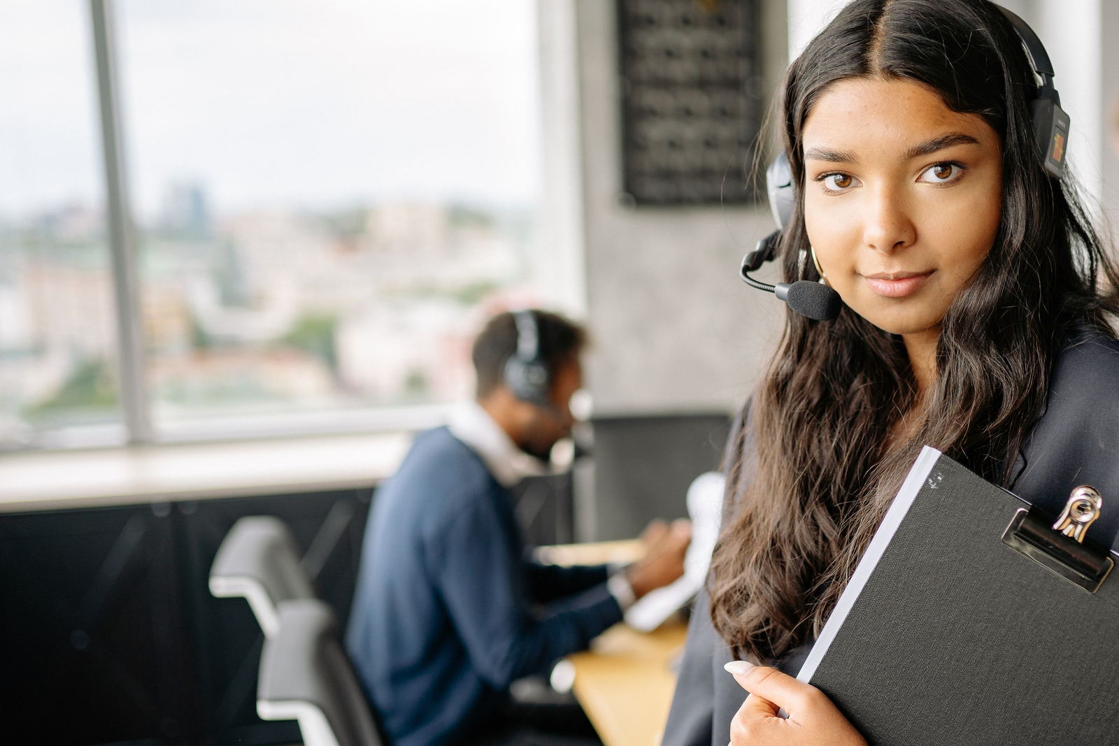 Support executive wearing a headset and holding a clipboard in a bright office, a colleague working behind her.