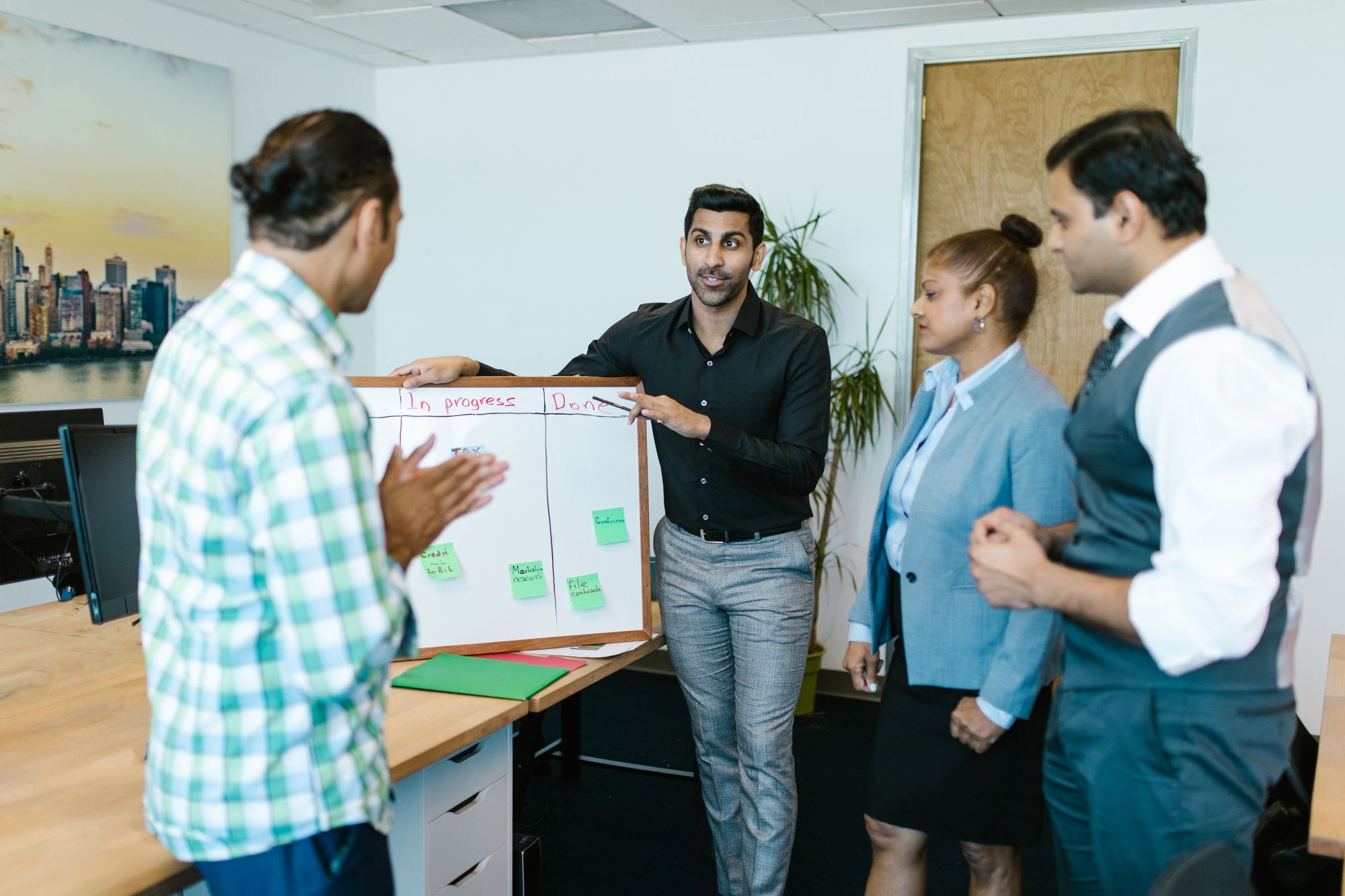 A team of four standing at a whiteboard in a daylit office, planning work together