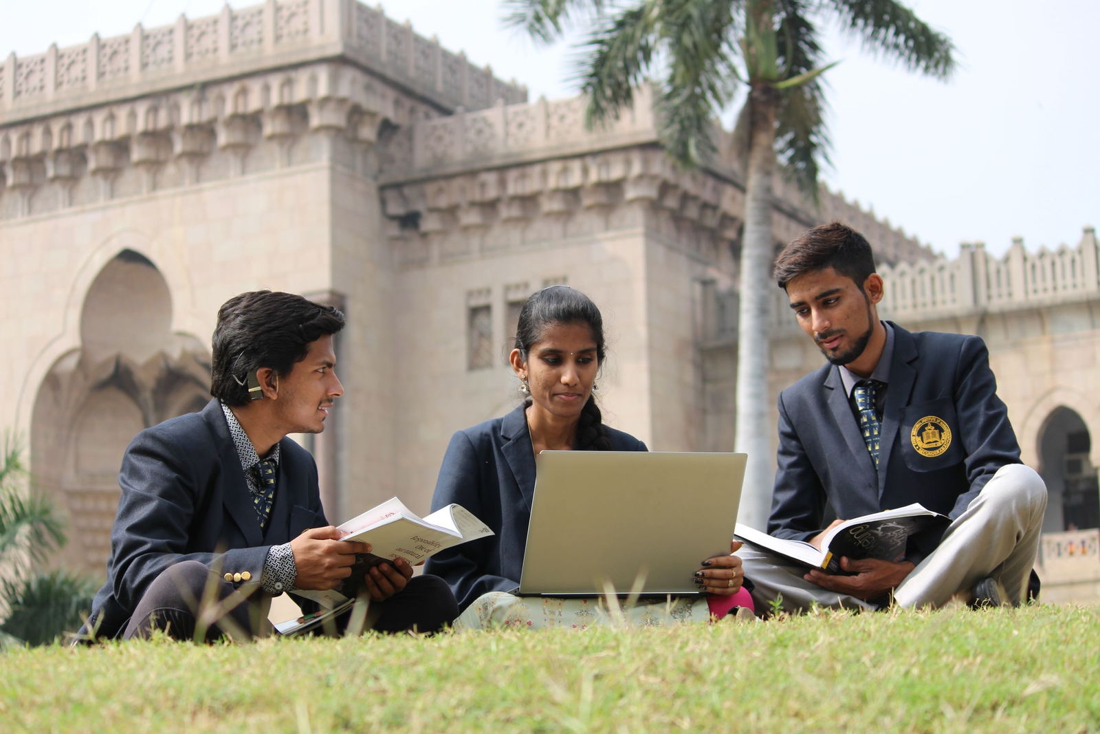 Three students in college blazers sharing a laptop and textbooks on a campus lawn